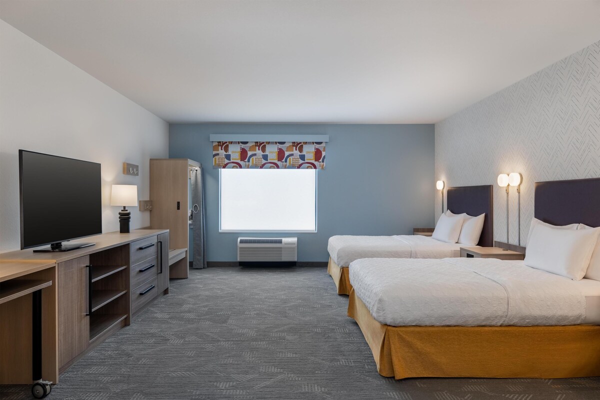 Hotelkamer met twee bedden, een bureau met een televisie, een kledingkast en een raam met een gestreept gordijn.