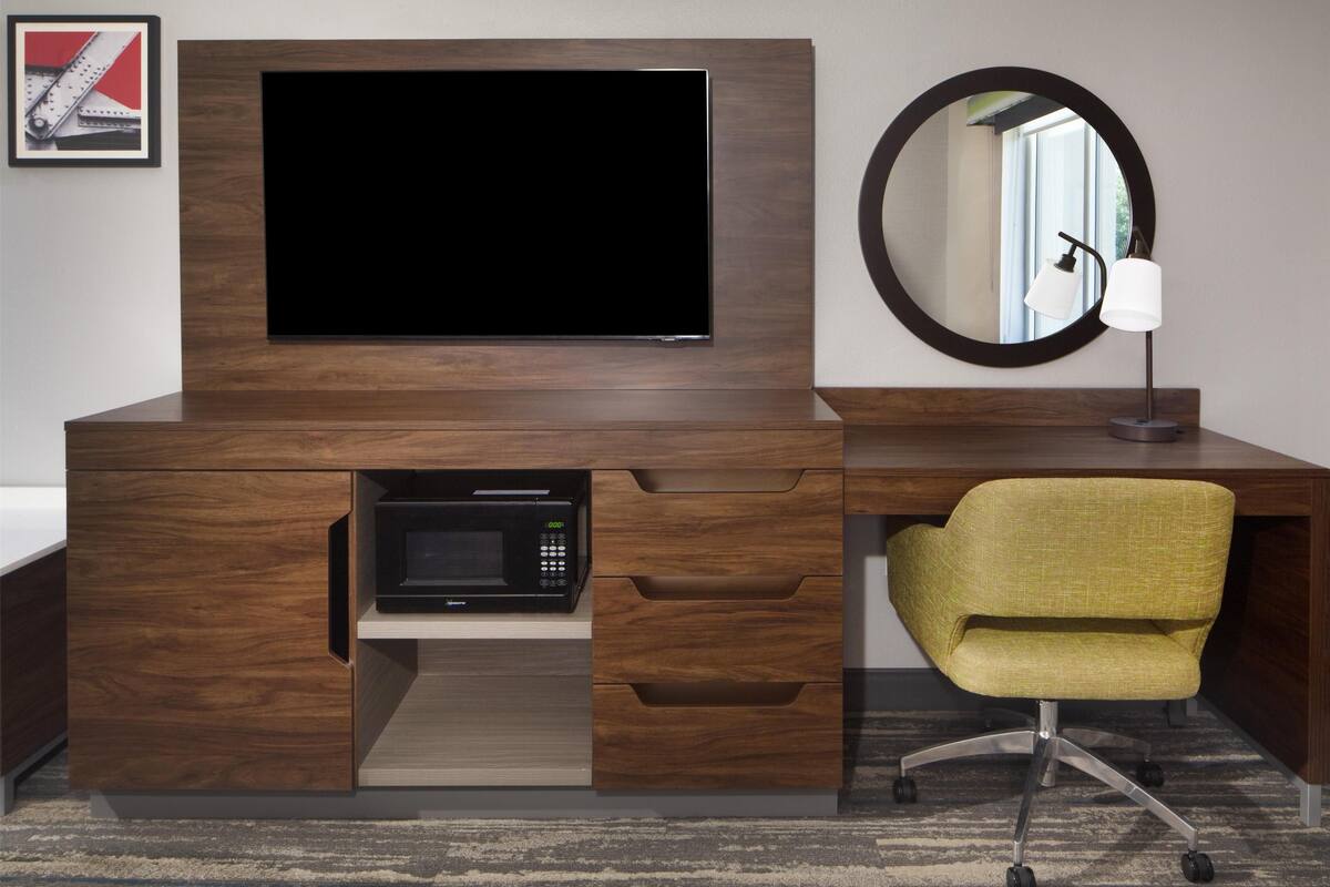 A modern hotel room with a wooden desk, a flat-screen TV, a microwave, a yellow chair, and a round mirror on the wall.