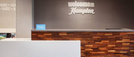En receptionsdisk med et 'Welcome to Hampton' skilt, et 'Hilton Honors' skilt, og en væg hvor ordet 'Treats' er gentaget.