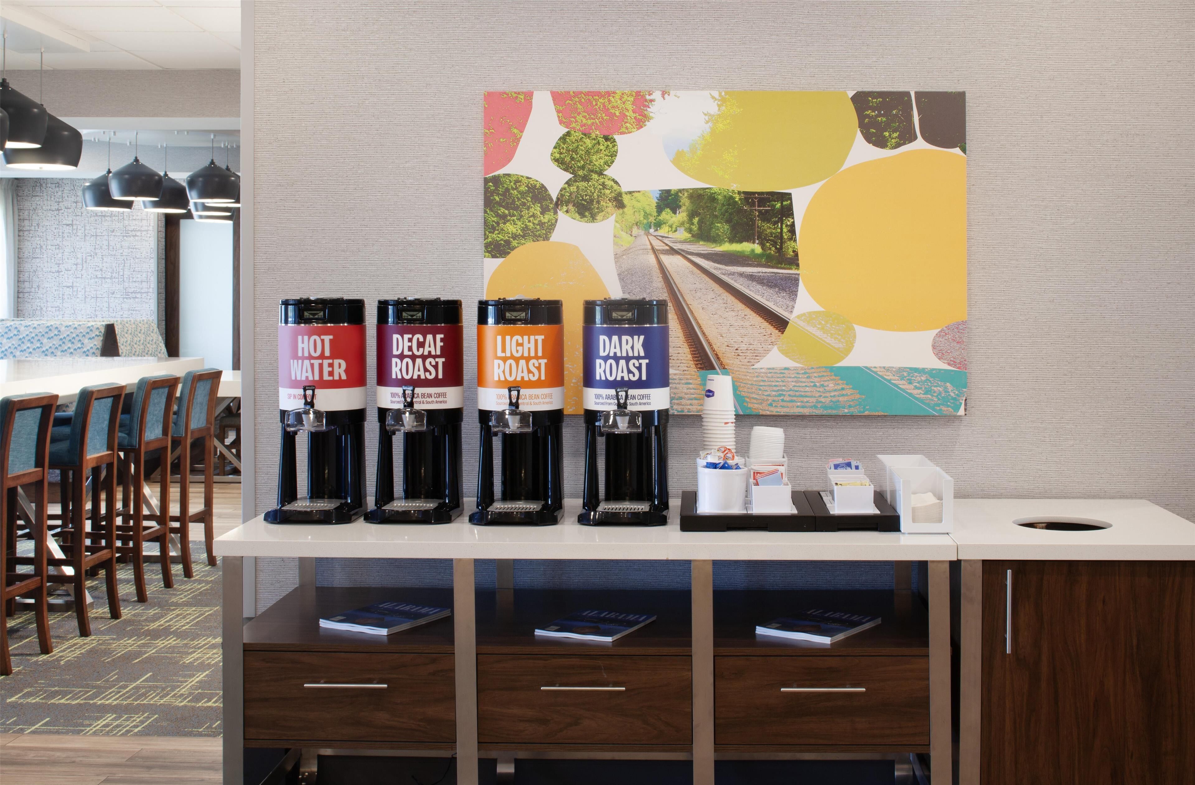 Un espace dédié au café, équipé de quatre distributeurs de café étiquetés « Eau chaude », « Café décaféiné », « Café léger » et « Café corsé ». On y trouve également des serviettes en papier et une petite poubelle sur le comptoir.