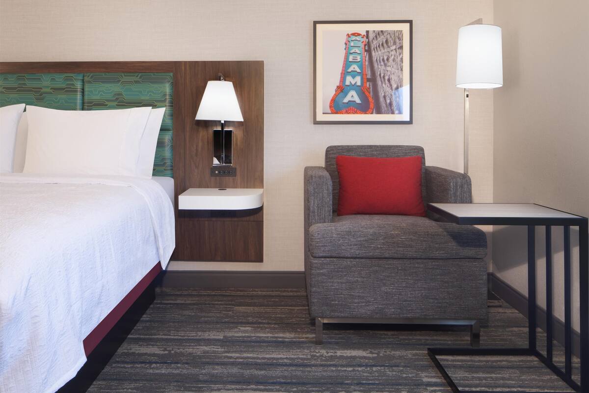 A hotel room with a bed, a desk, a chair, a nightstand, a lamp, and a framed picture on the wall.