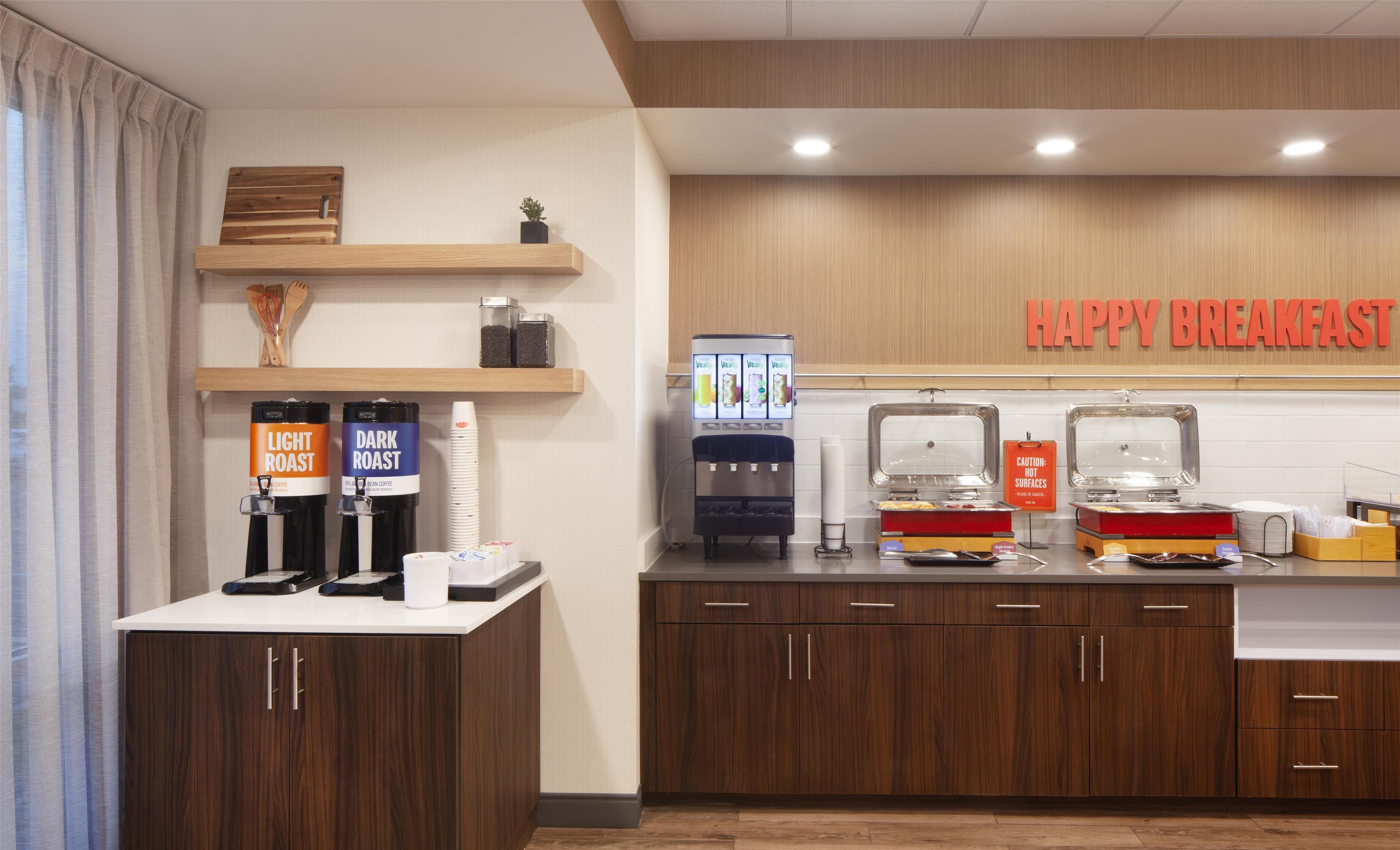 A breakfast area with coffee dispensers, a beverage vending machine, and a sign that reads "HAPPY BREAKFAST".
