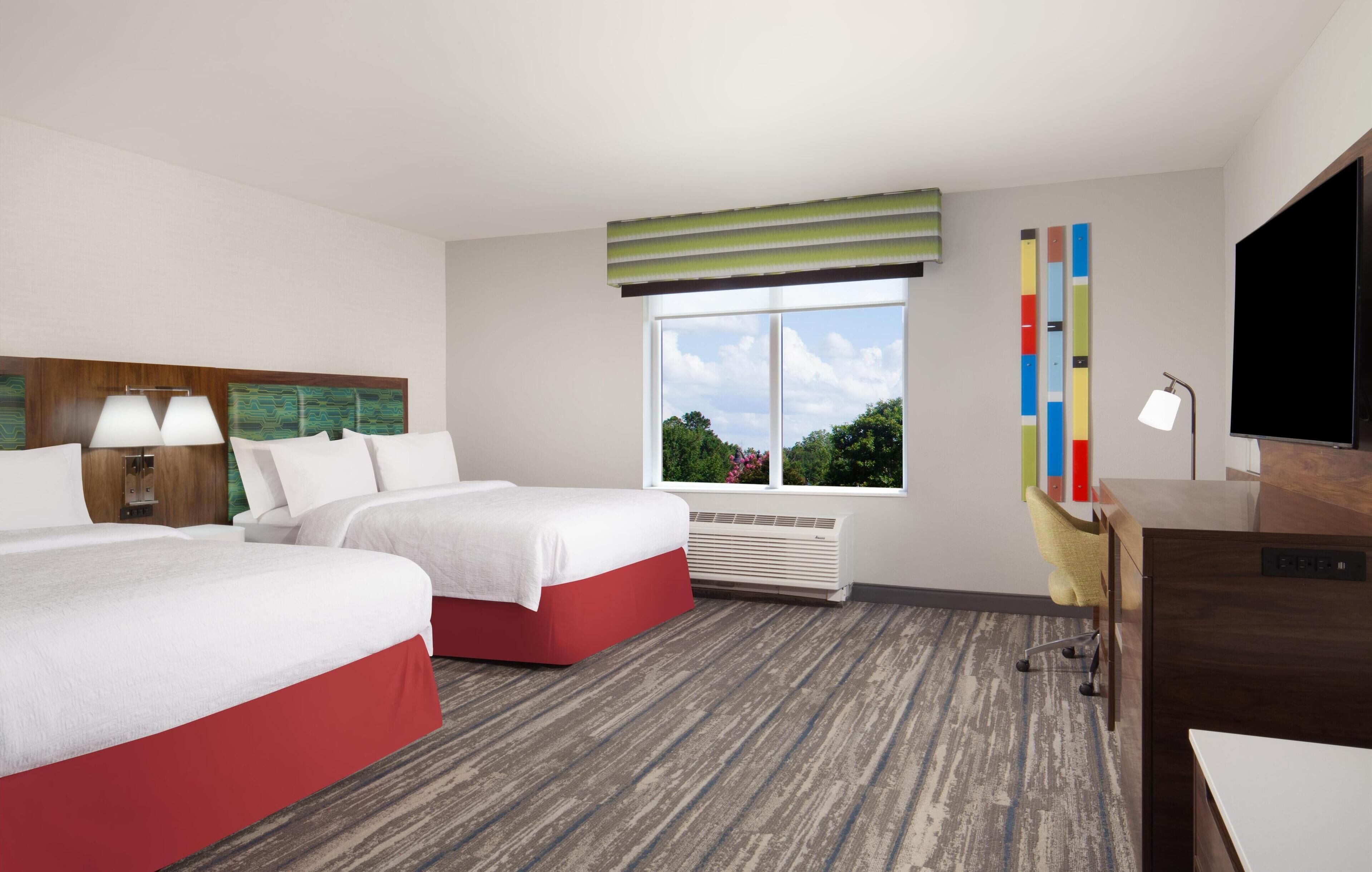 A hotel room with two beds, a desk, a TV, and a window with a view of trees.