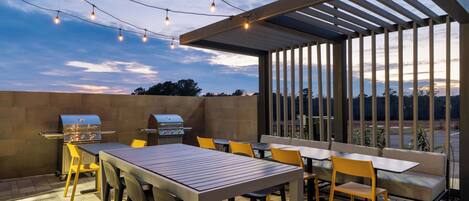 An outdoor patio with a grill, tables, and chairs, under a pergola with string lights.