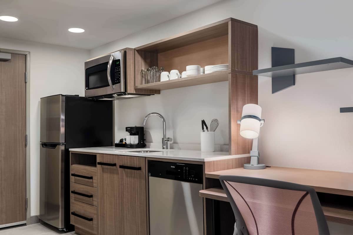 Una cocina moderna con microondas integrado, cafetera y lavavajillas. Hay un estante de madera sobre el fregadero con tazas y vasos.