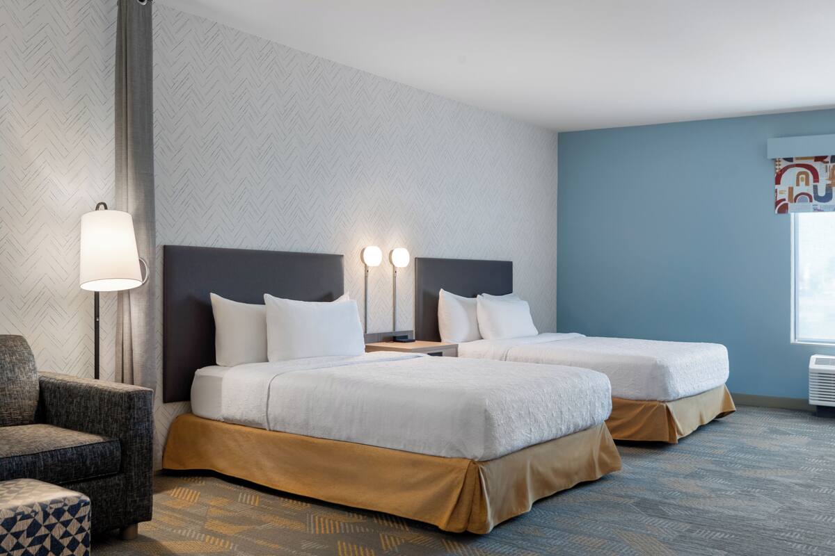 Habitación de hotel con dos camas, una mesita de noche, una lámpara, una silla y una alfombra estampada.