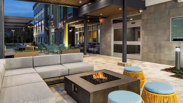 An outdoor seating area with a fire pit, cushioned seating, and a view of the building's interior. - Home2 Suites By Hilton Troy, Al (Troy)