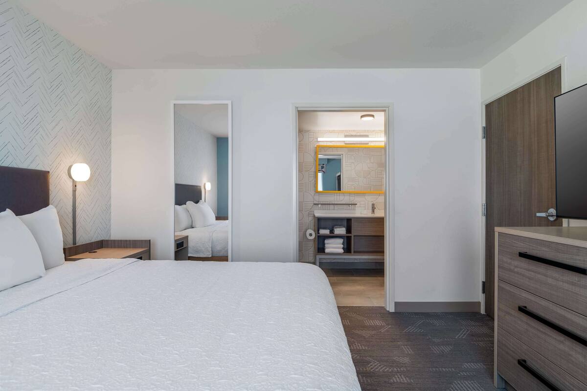 Habitación de hotel con una cama grande, una lámpara de noche, un armario y un baño visible a través de una puerta abierta.