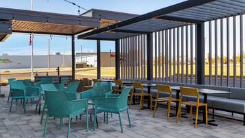 An outdoor seating area with tables and chairs, a covered section, and a view of a commercial building in the background.