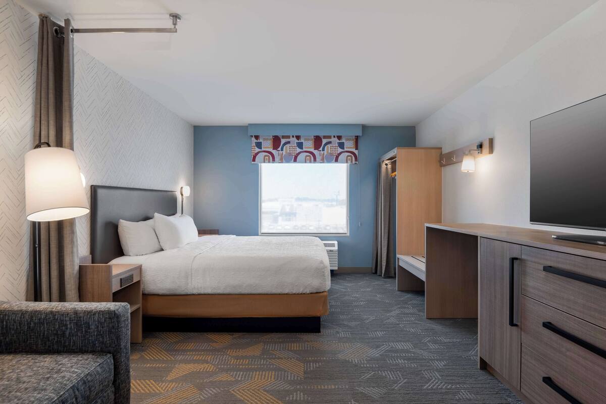 Habitación de hotel con cama, mesita de noche, lámpara, televisor, armario y vistas a la ciudad a través de la ventana.