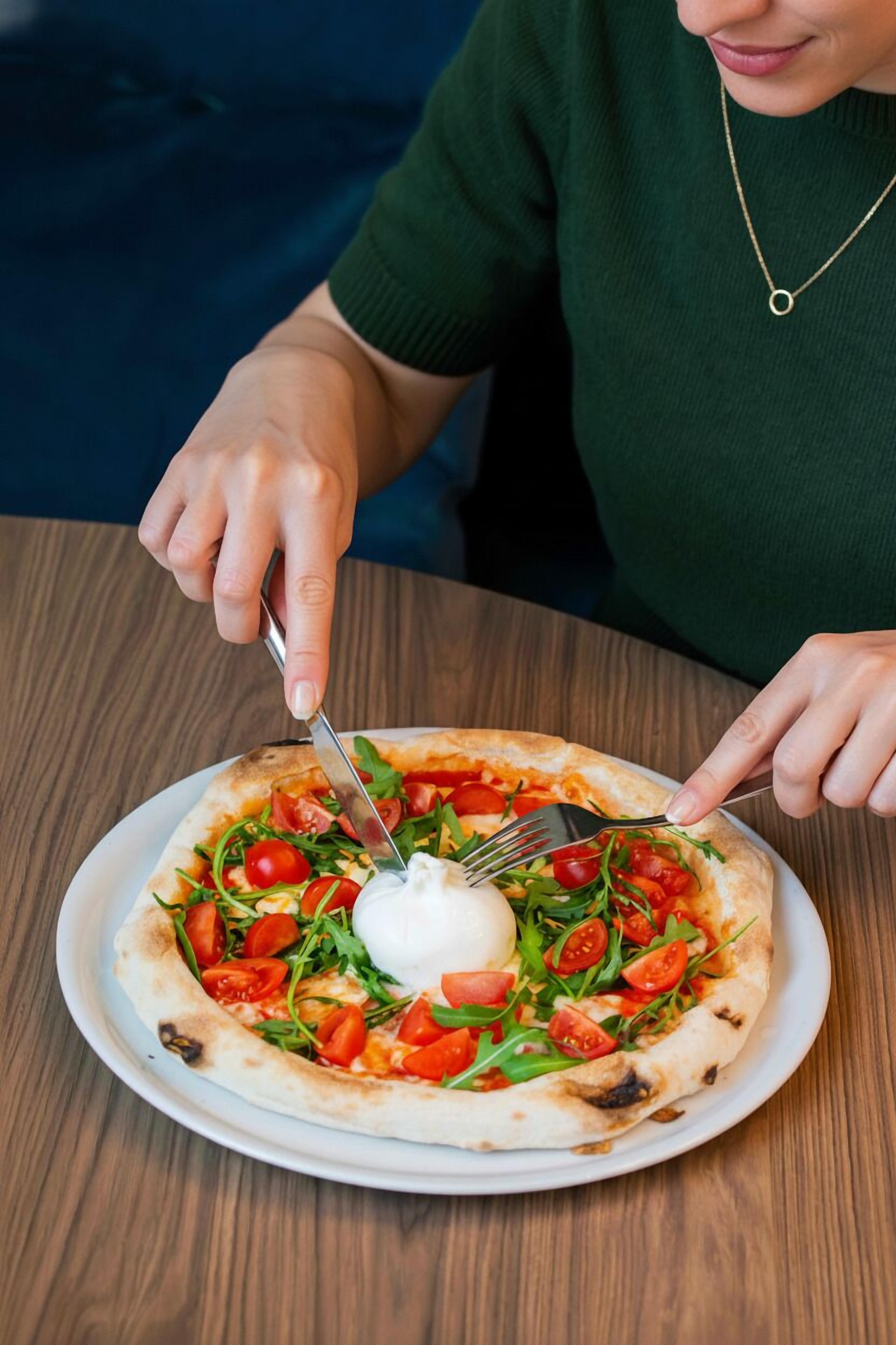 A person cutting a pizza with a fork and knife on a wooden table.