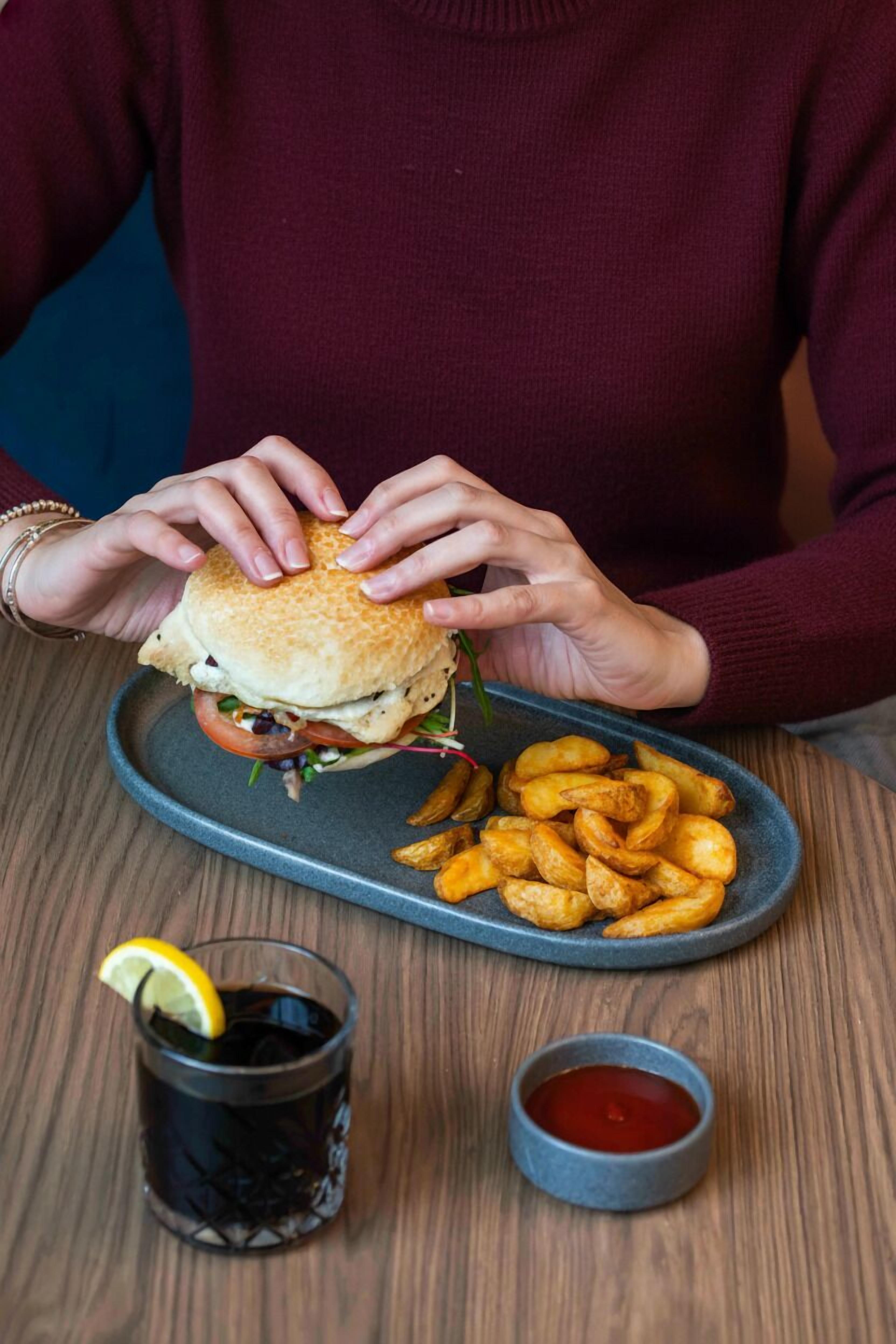 A person holding a burger with fries and a drink on a tray.
