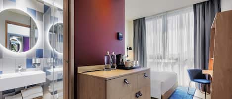 A modern hotel room with a large bed, a wooden dresser, a bathroom with a glass shower enclosure, and a view of the city through the window.