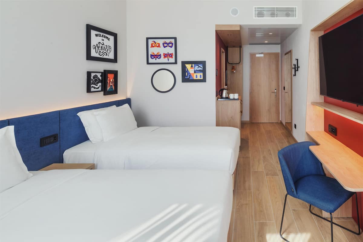 Habitación de hotel con dos camas, un escritorio, una silla y un televisor. Se observan cuadros en la pared y una puerta que da a una kitchenette.