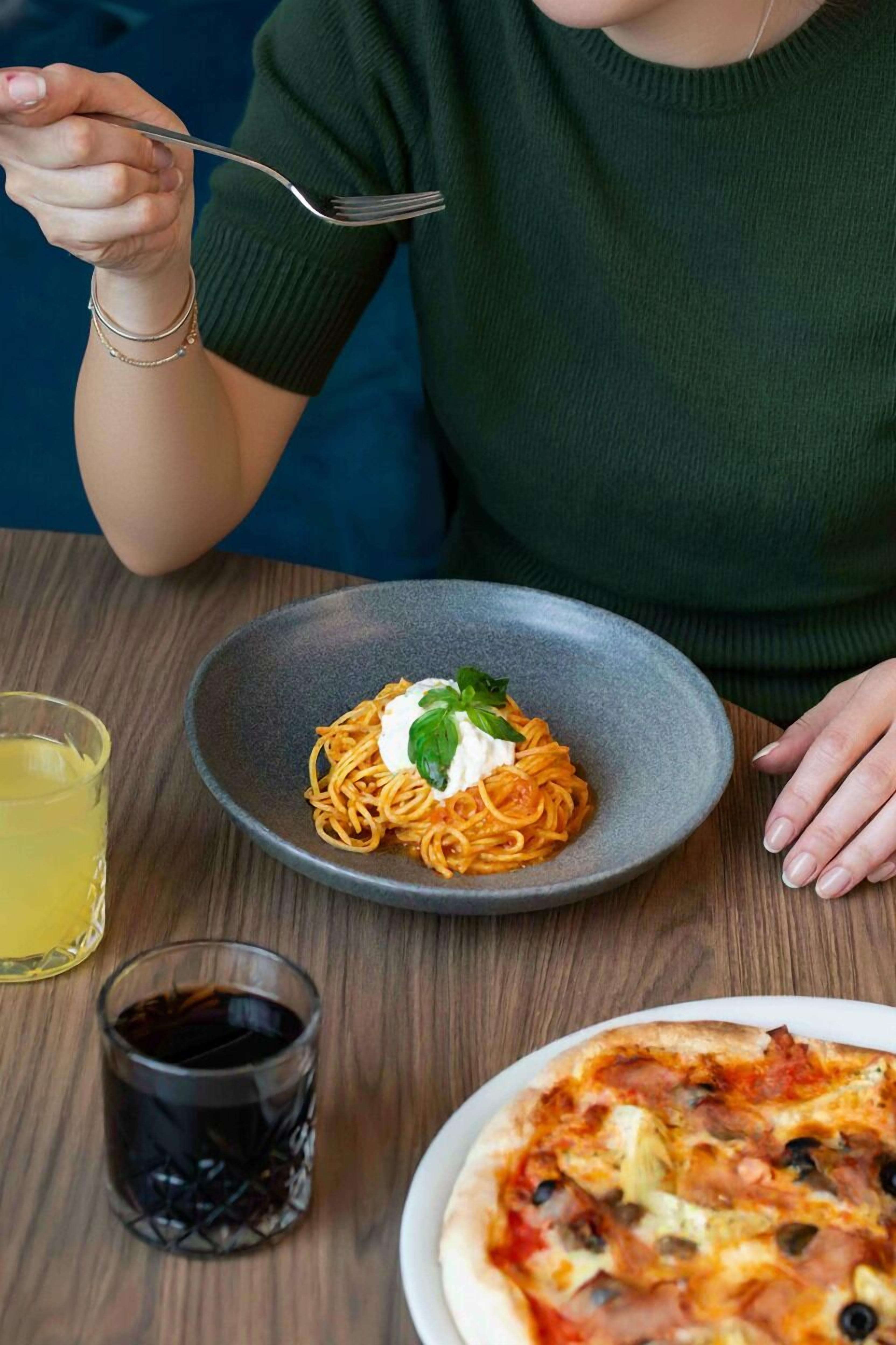 Una persona cenando un plato de espagueti, un vaso de refresco y una pizza sobre una mesa de madera.