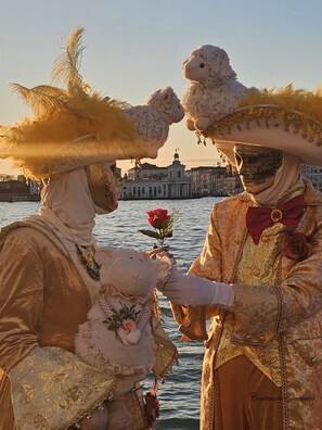 Duas pessoas em trajes históricos perto de um corpo d'água, com um edifício ao fundo.