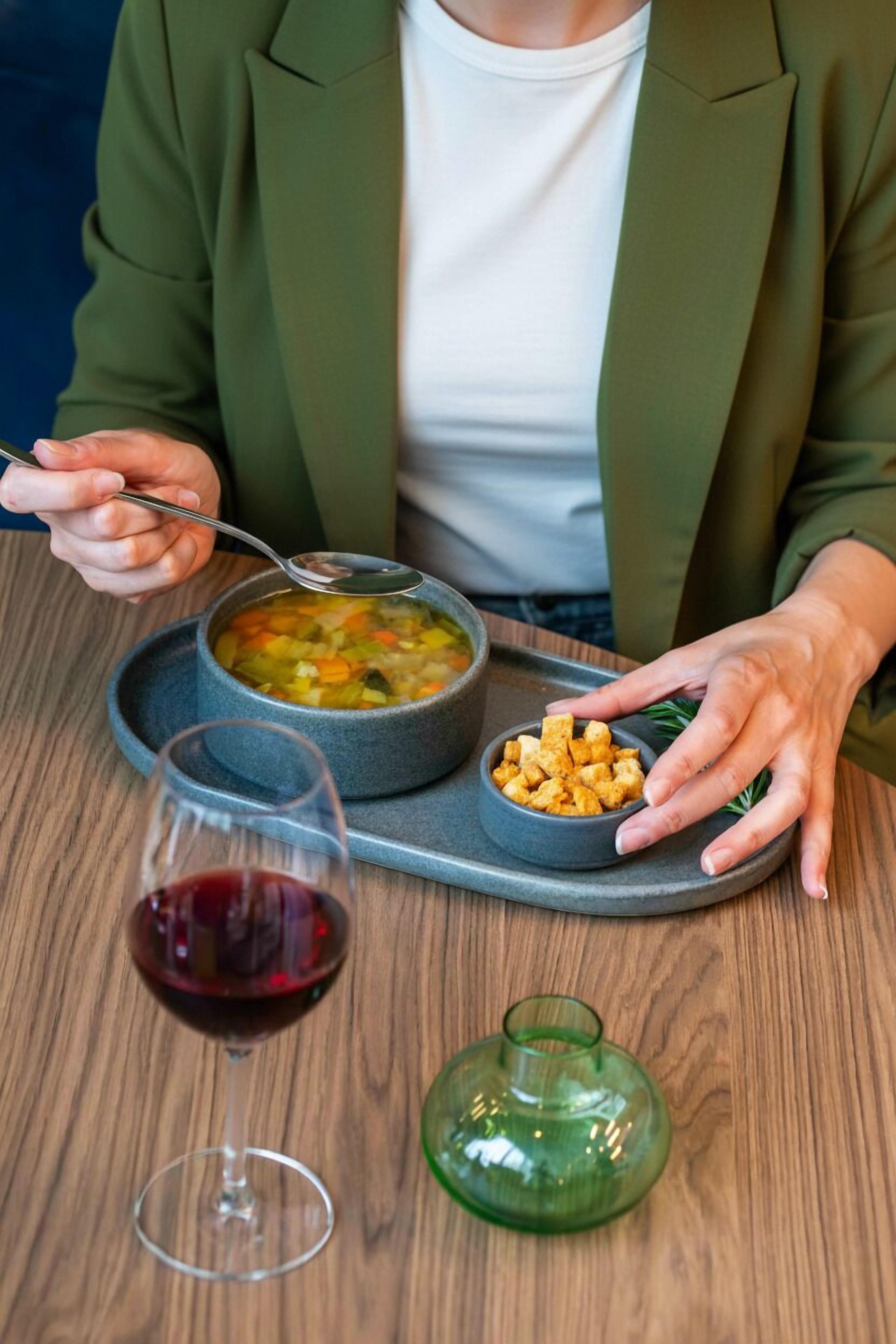 Eine Person sitzt an einem Tisch mit einer Suppenschüssel, einem Glas Rotwein und einer kleinen Schale Croutons.