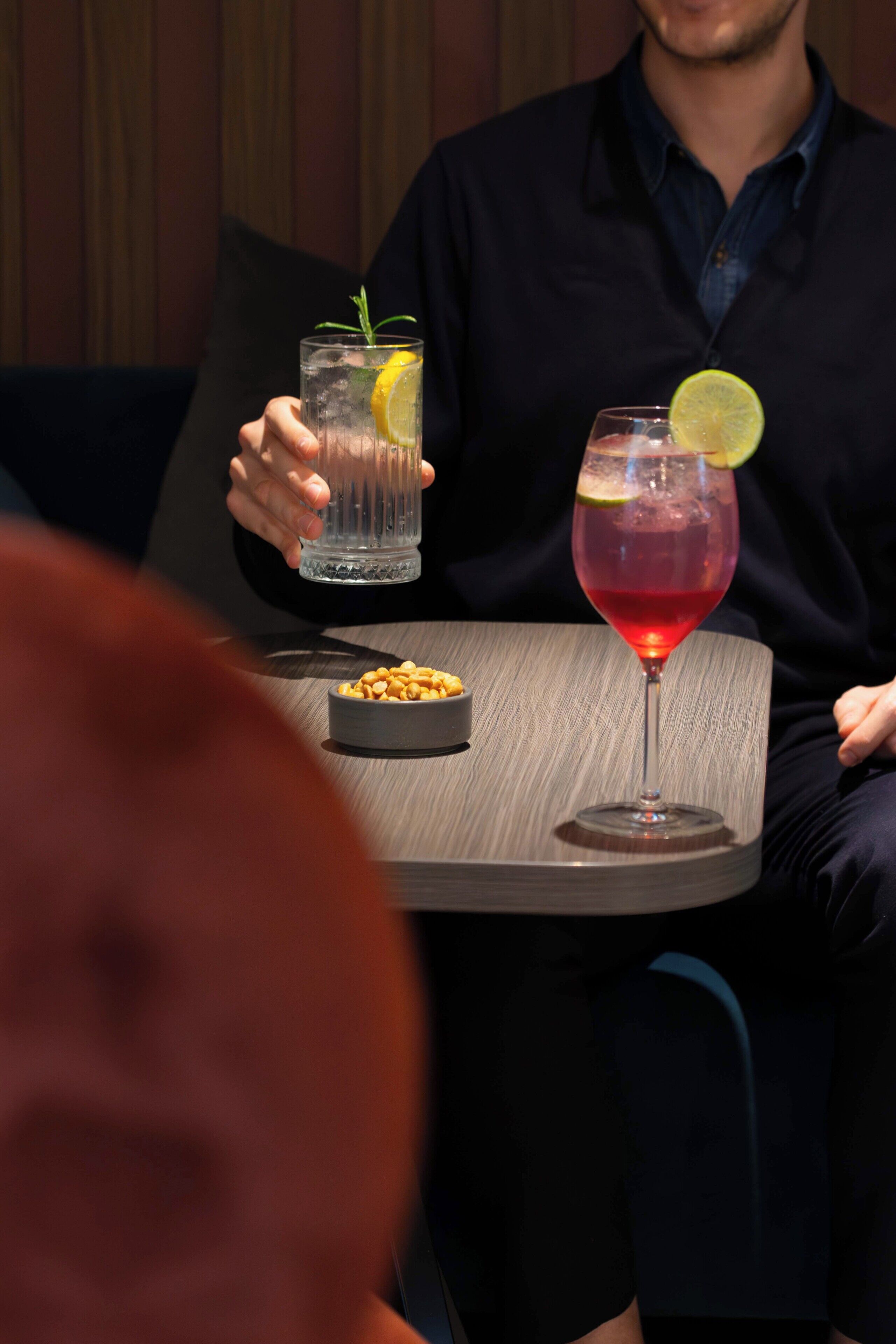 A person holding two drinks, one in a glass and one in a tumbler, with a plate of nuts on the table.