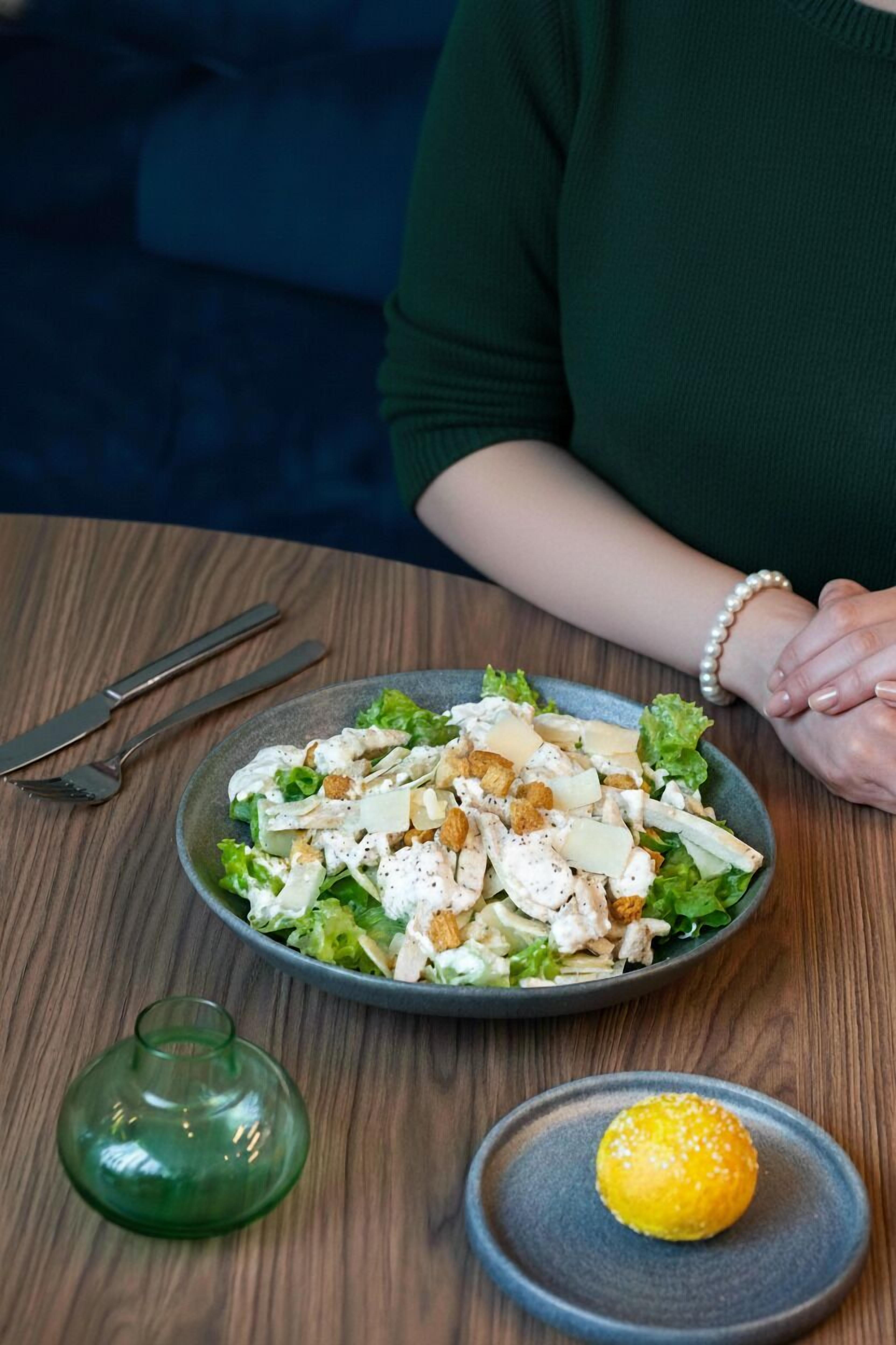 Una persona sentada en una mesa con una ensalada, un vaso verde y un plato pequeño con algo amarillo.