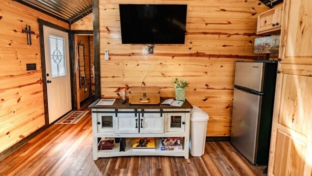 A compact kitchen with wooden walls, a flat-screen TV mounted above the counter, and a refrigerator to the right.