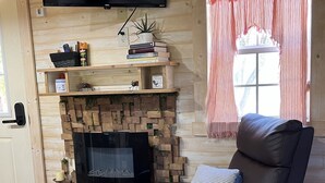 A cozy living room with a fireplace, a flat-screen TV mounted above it, and a comfortable leather sofa. - Tiny Farmhouse Pickleball Court and Pet Friendly! (Granbury)