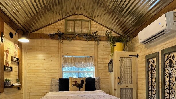 A cozy wooden room with a bed, a ceiling light fixture, and an air conditioning unit. - Tiny Farmhouse Pickleball Court and Pet Friendly! (Granbury)