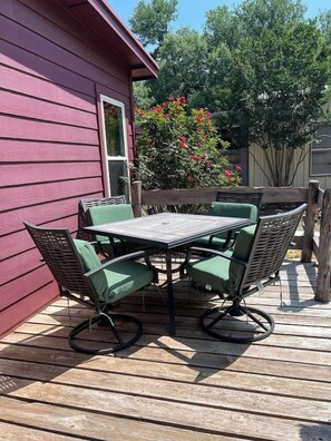 Une terrasse en bois avec une table et des chaises, un bâtiment rouge et un arbuste en fleurs.