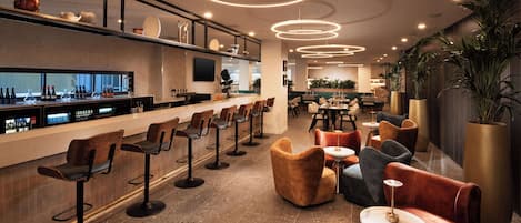 A modern bar area with a long counter, multiple bar stools, and a mix of seating options including sofas and round tables.