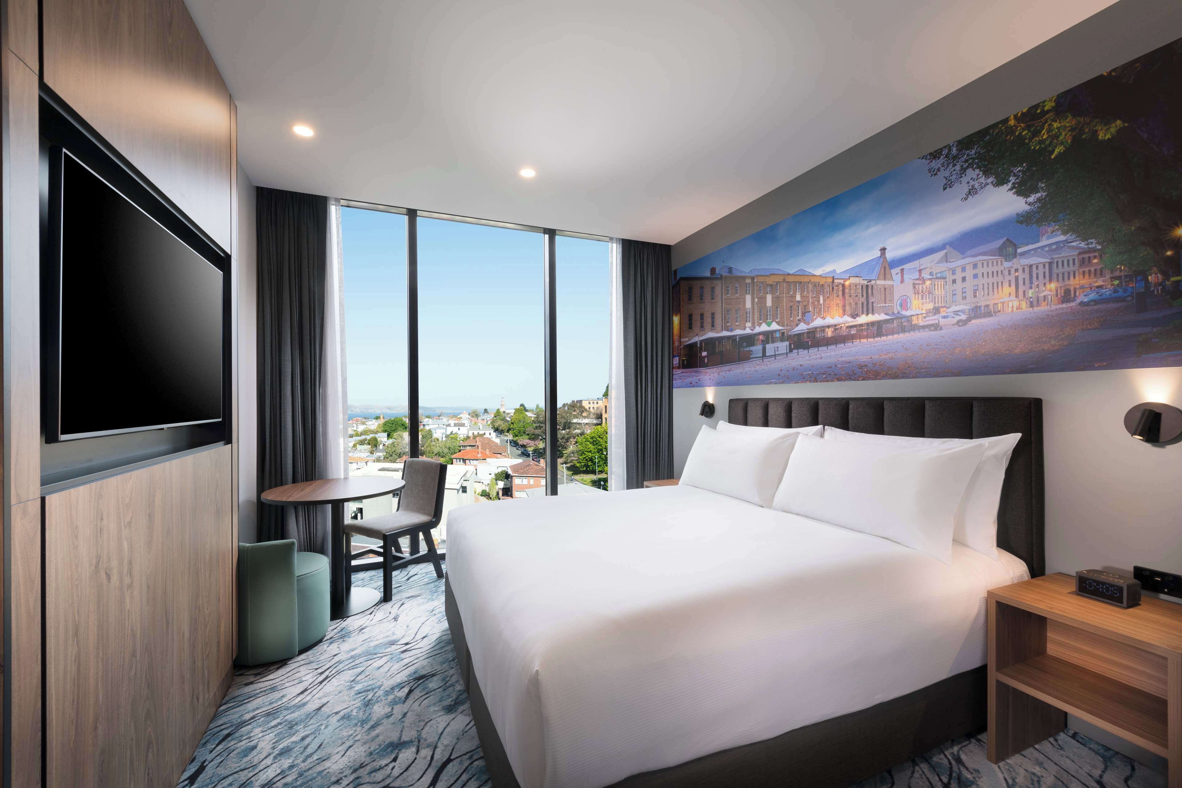 A hotel room with a large bed, a flat-screen TV mounted on the wall, a small table and chair, and a painting of a cityscape on the wall.