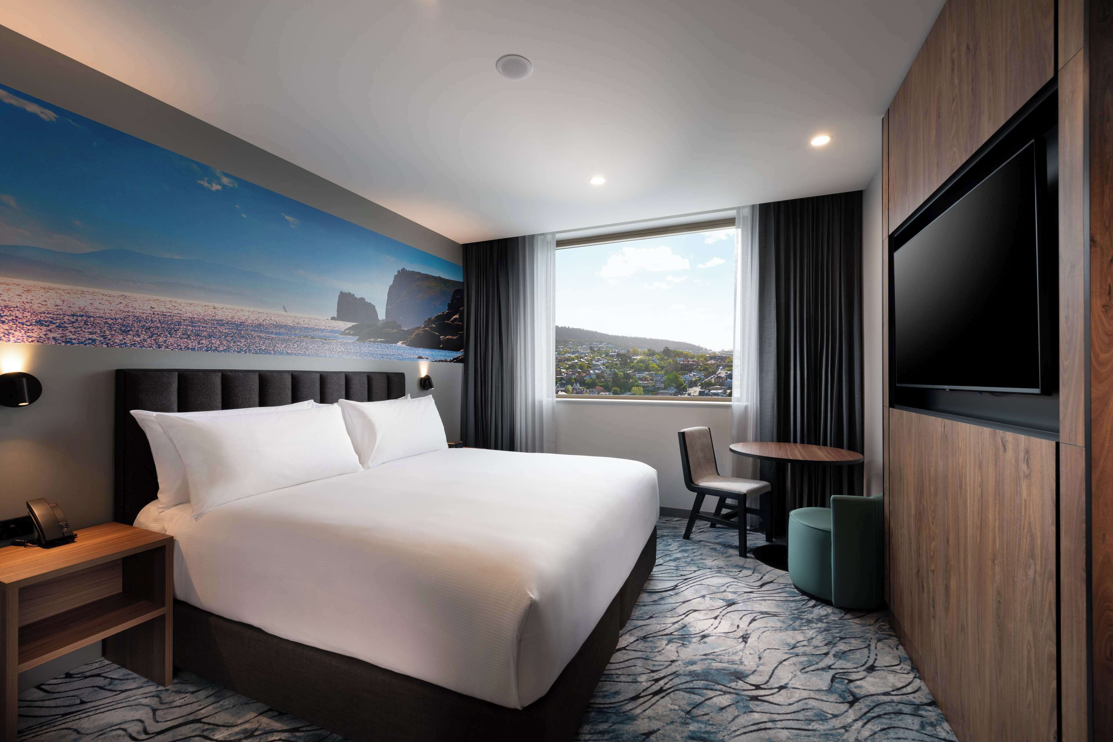 A hotel room with a large bed, a TV, a desk with a chair, and a view of the city through a large window.