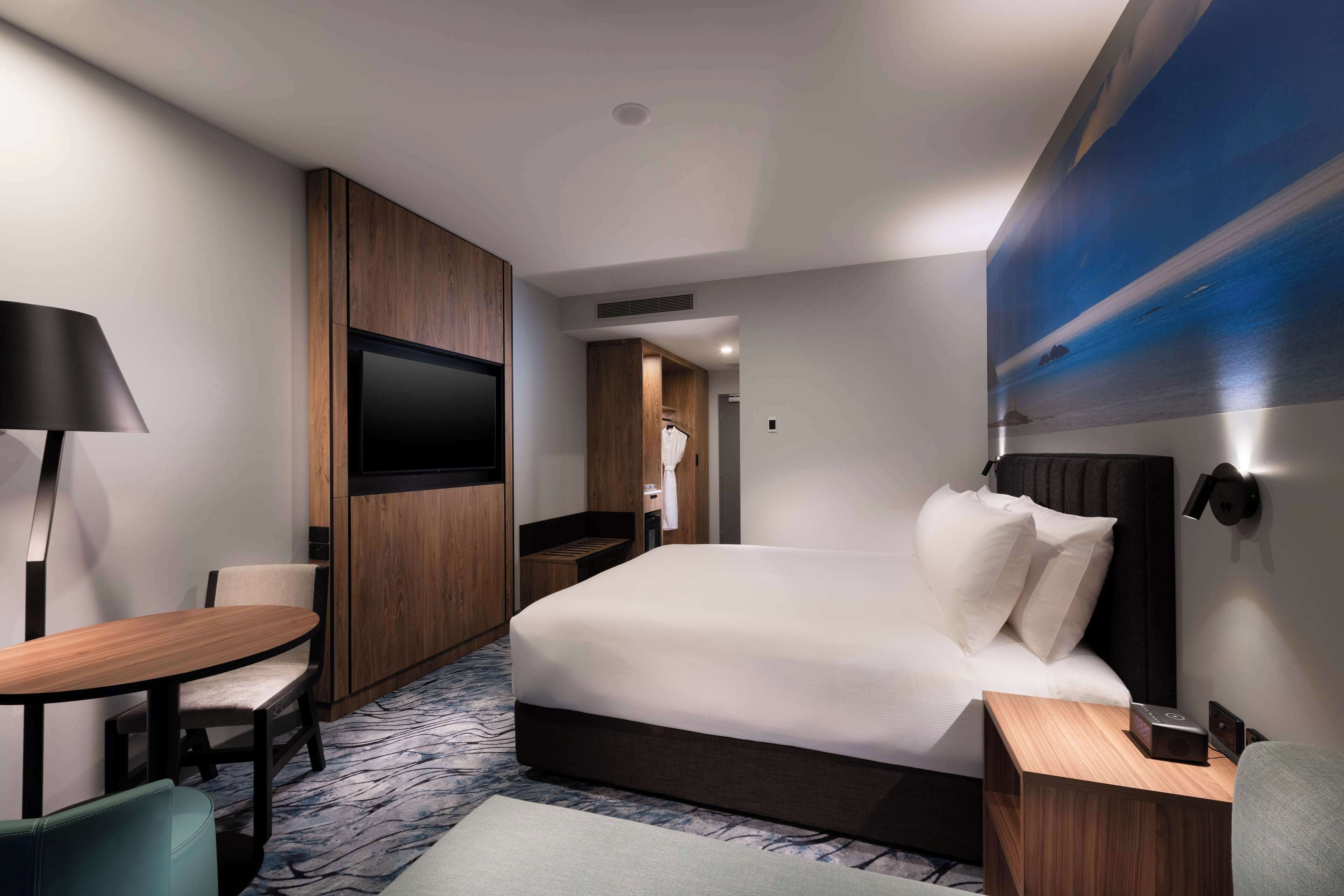 A modern hotel room with a large bed, a desk with a chair, a flat-screen TV, and a wall-mounted light.