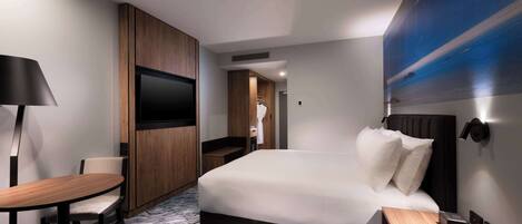 A modern hotel room with a large bed, a desk with a chair, a flat-screen TV, and a wall-mounted light.