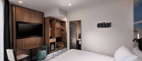 A modern hotel room with a large bed, a flat-screen TV mounted on a wooden panel, a small round table with a chair, and a walk-in closet with a mirror.