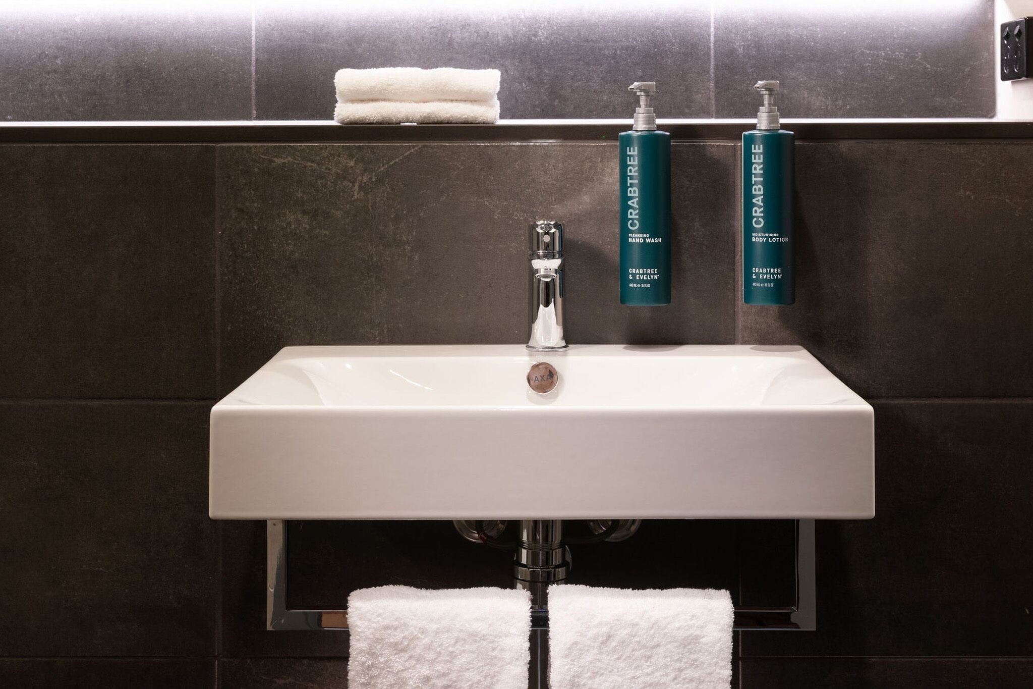 Un baño con un lavabo blanco, dos botellas de loción corporal Crabtree y un perchero con dos toallas.