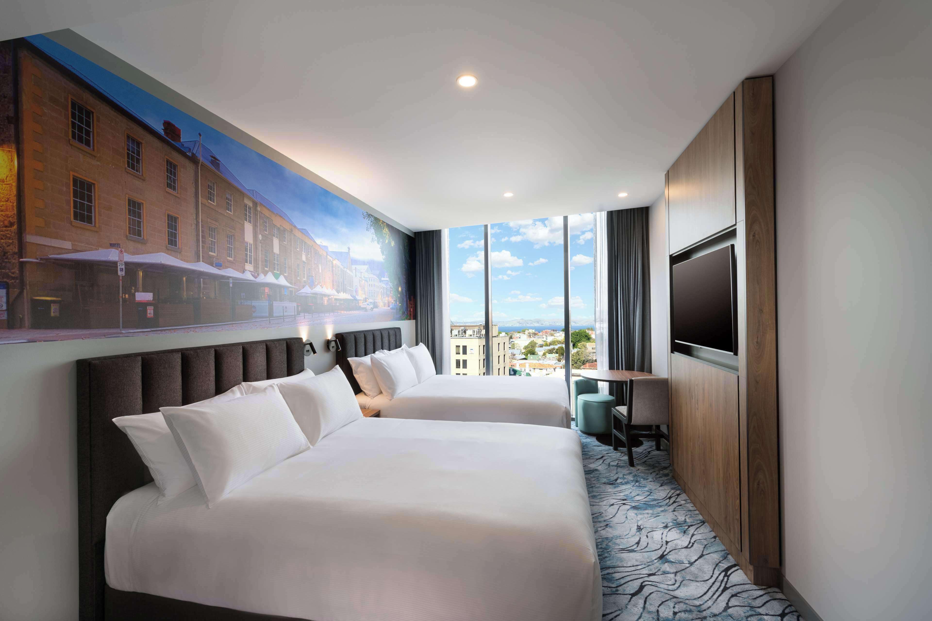 Habitación de hotel con dos camas, un televisor grande y vistas a la ciudad.