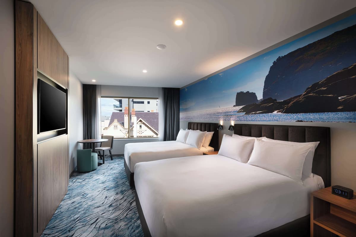 A hotel room with two beds, a TV, a desk, and a large window with a view of buildings.