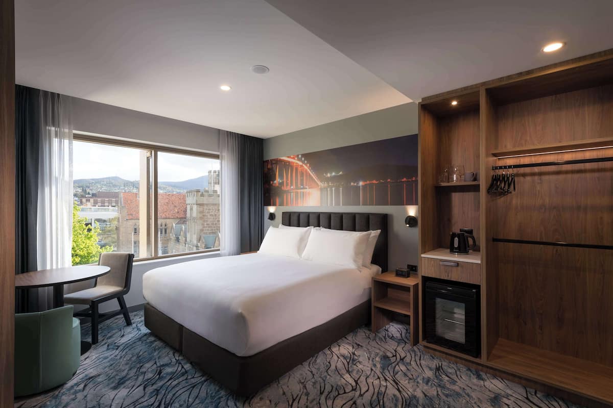 A hotel room with a large bed, a desk, a chair, a small refrigerator, and a view of the city.