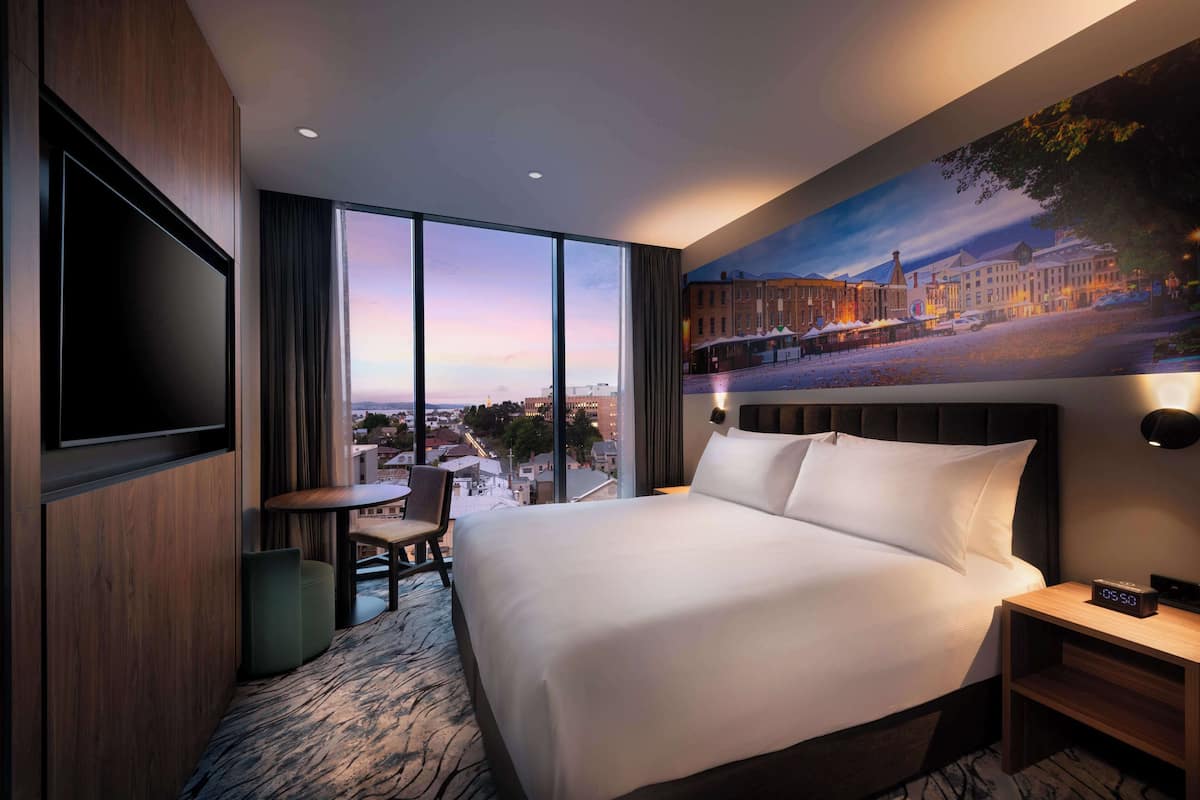A hotel room with a large bed, a TV, a small table, and a chair. There is a painting of a cityscape on the wall.