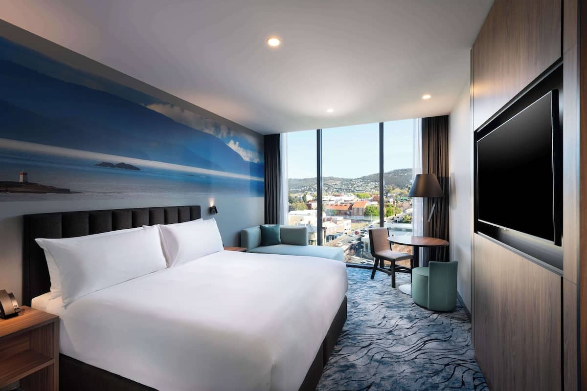 A hotel room with a large bed, a seating area with a table, a TV, and a view of the city and hills.