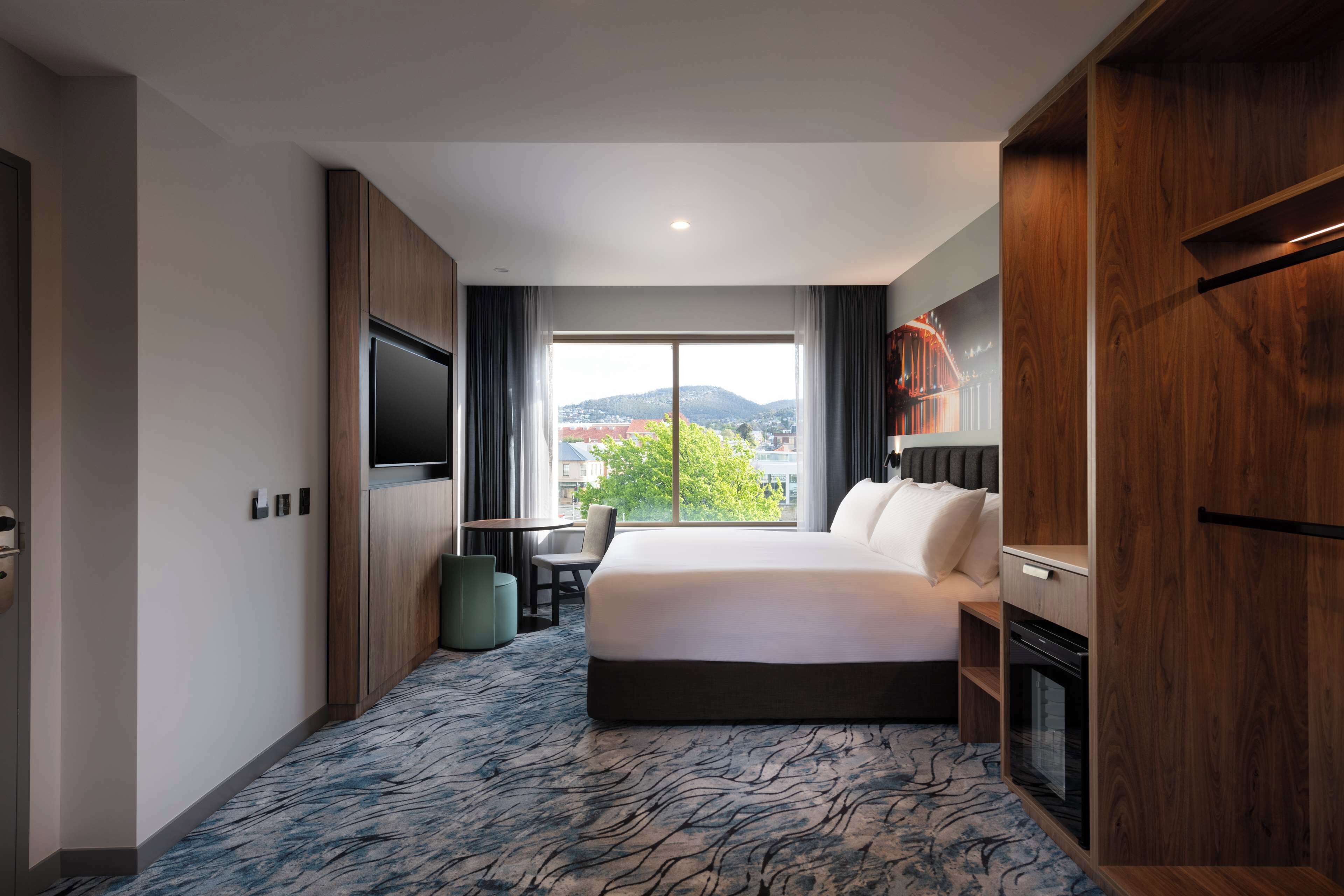 A hotel room with a large bed, a flat-screen TV, a dining table, and a view of the cityscape.