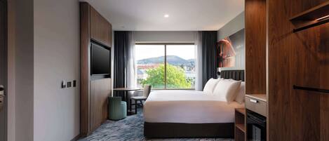 A hotel room with a large bed, a flat-screen TV, a dining table, and a view of the cityscape.
