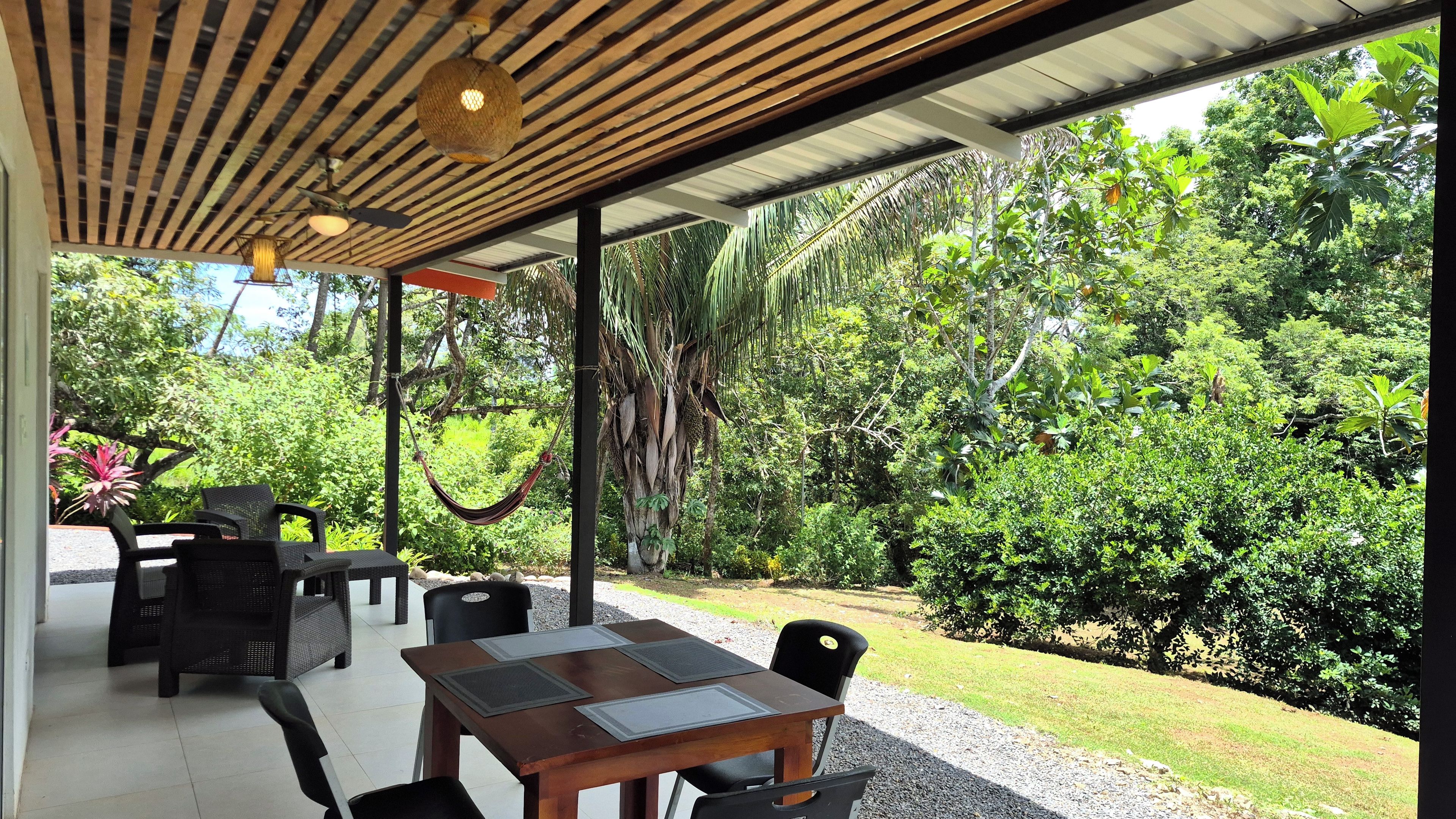 An outdoor patio with a wooden table and chairs, surrounded by lush greenery and a hammock.