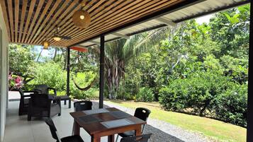 An outdoor patio with a wooden table and chairs, surrounded by lush greenery and a hammock.