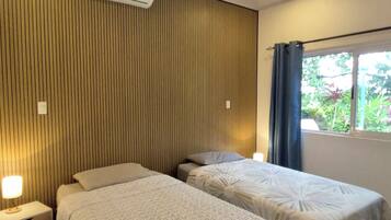 A hotel room with two single beds, a ceiling fan, an air conditioning unit, a window with curtains, and a bedside table with a lamp.