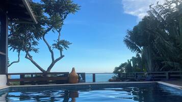 A swimming pool with a view of the sea, surrounded by tropical trees and a bench.