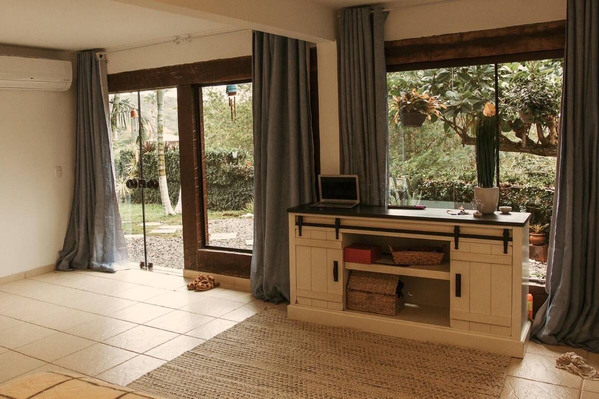 A room with a desk, a laptop, and a view of a garden through large windows.