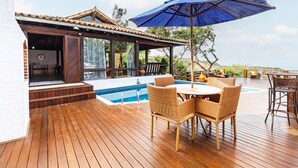 A wooden deck with a pool, outdoor seating, and a covered area with a blue umbrella. - Maravilha Pousada (Búzios)