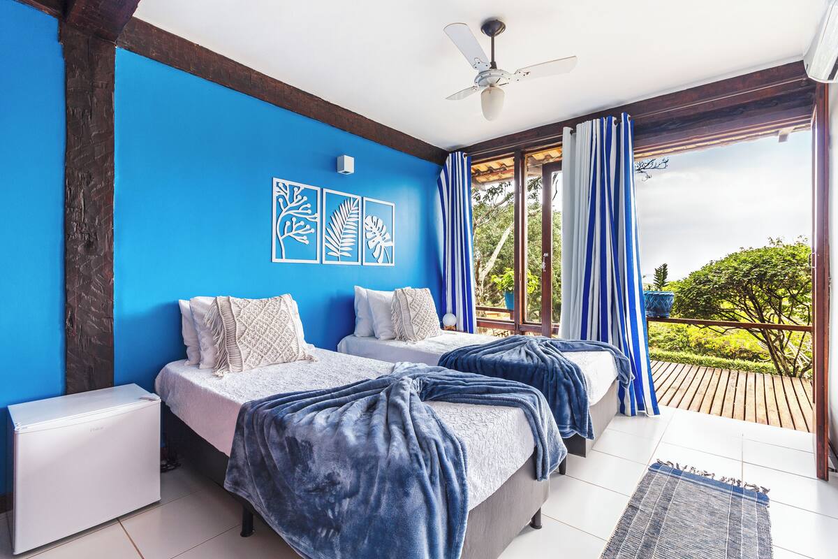 A bedroom with two beds, a ceiling fan, and a view of a wooden deck and greenery.