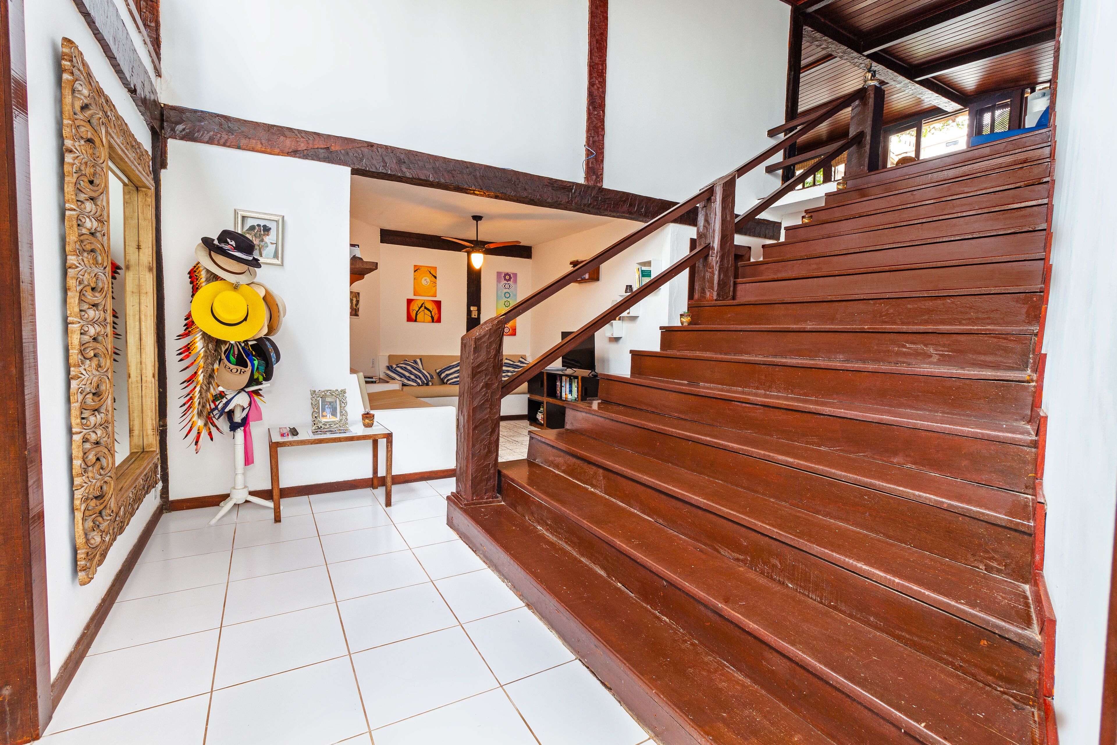 A wooden staircase with a handrail leading to an upper floor. A mirror with an ornate frame is on the left, and a wall with framed pictures and a small table with a picture frame and a vase is visible.