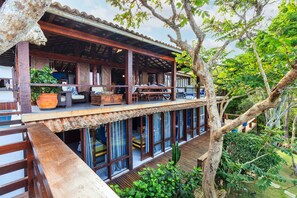 A two-story wooden structure with a balcony, large windows, and a view of the surrounding greenery.